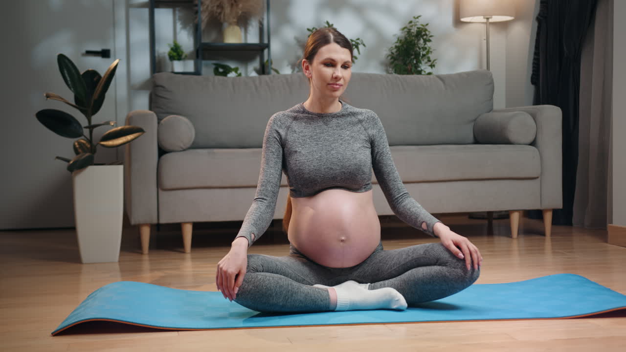 Pregnant woman in sportswear performs seated spinal twist on yoga mat in living room promoting