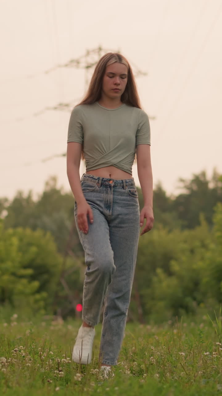 Young woman walking along grassy path with power lines overhead and blurred background, featuring distant red light and passing car, she moves thoughtfully through serene, green surroundings