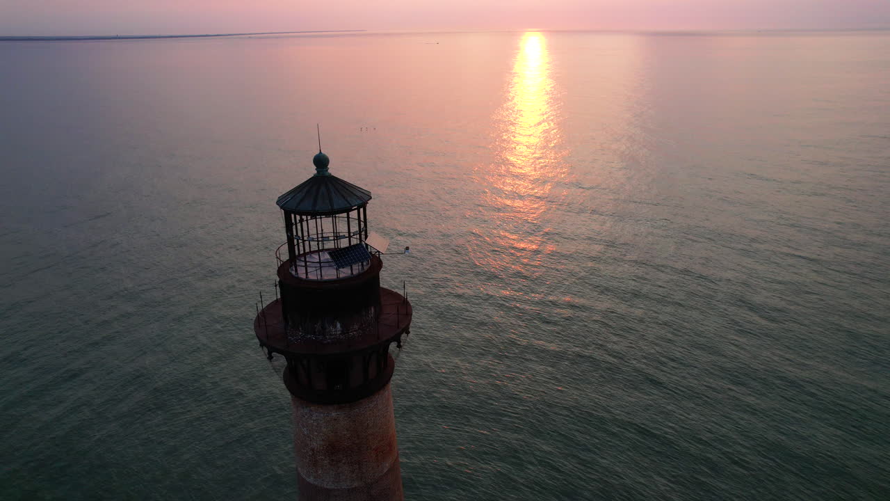 A drone shot moving down while angling up towards a lighthouse with the sunrise and ocean in the background.