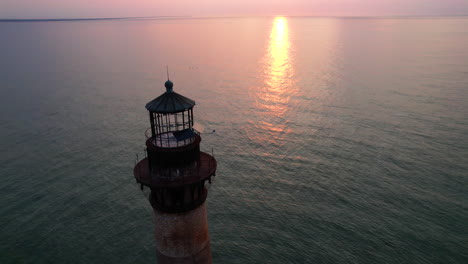a drone shot moving down while angling up towards a lighthouse with the sunrise and ocean in the background.