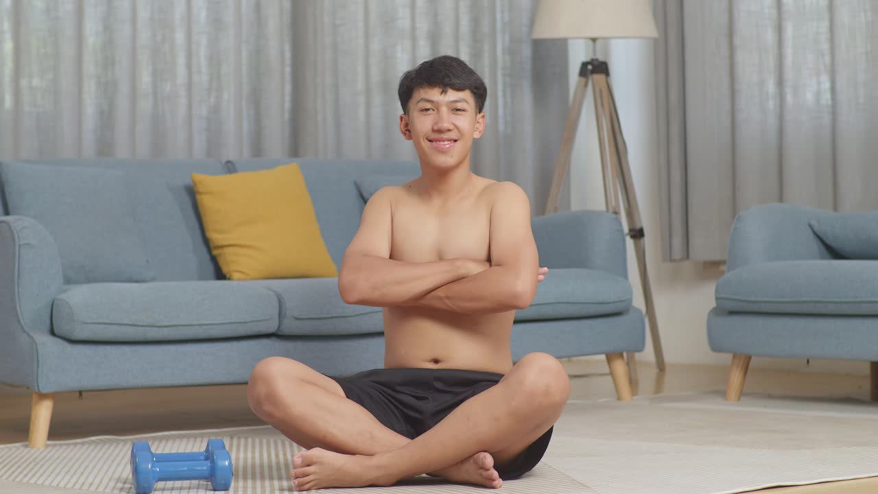 아시아 십대 소년 은 집 에서 셔츠 를 벗고 운동 한 후 덤벨 을 들고 매트 에 앉아 있는 동안 팔 을 고 카메라 에 미소 짓고 있다