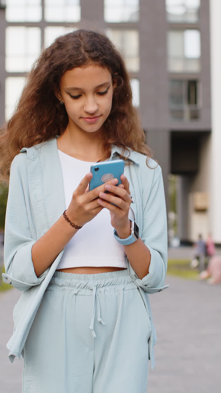chica preadolescente sonriente usando su teléfono inteligente escribiendo mensajes de texto respondiendo mensajes chateando en línea en la calle de la ciudad