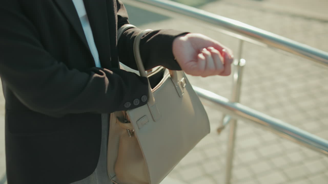 Close up of lady searching handbag outdoors, hand inside bag retrieving item, walking near metal railings, dressed in professional attire with sunlight casting shadows on pavement