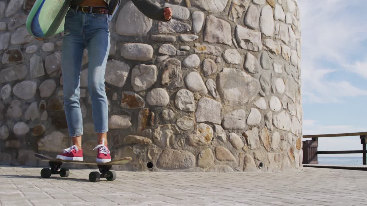 Glad kvinne av blandet etnisitet skateboarder, bærer surfebrett på en solrik promenade ved sjøen.