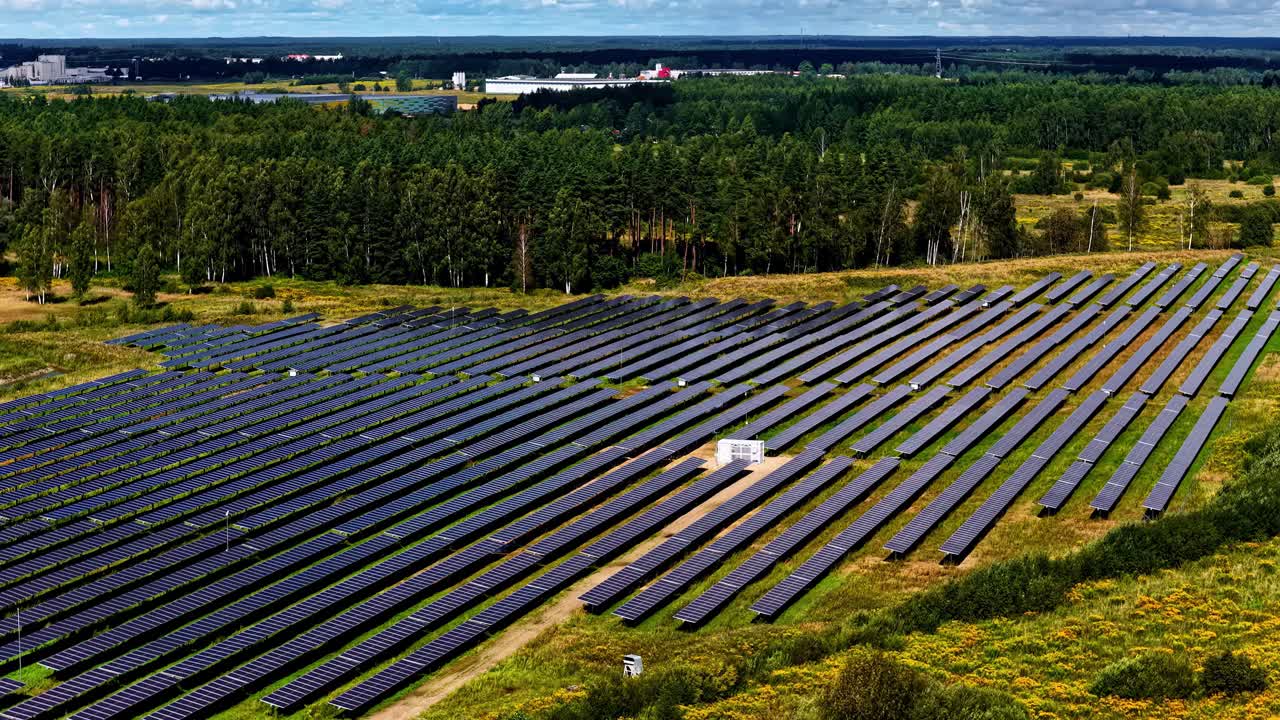 Luftfoto af en stor solcellepark ved siden af skov og åbent land, der viser rækker af solcellepaneler, som producerer vedvarende energi i et landligt område