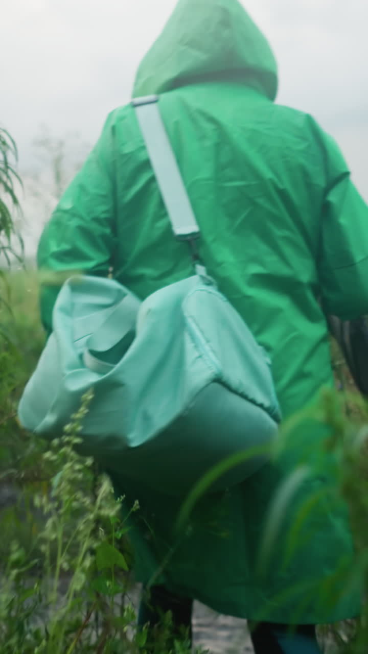 vista trasera de un individuo con un impermeable verde que lleva una bolsa de color menta y un paraguas, con botas de lluvia amarillas, caminando cuidadosamente por un camino rodeado de vegetación alta y plantas