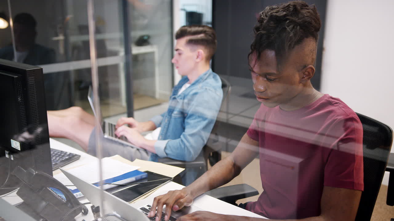 Elevated view of two young men working at computers sitting in a glass office cubicle, close up, seen through glass