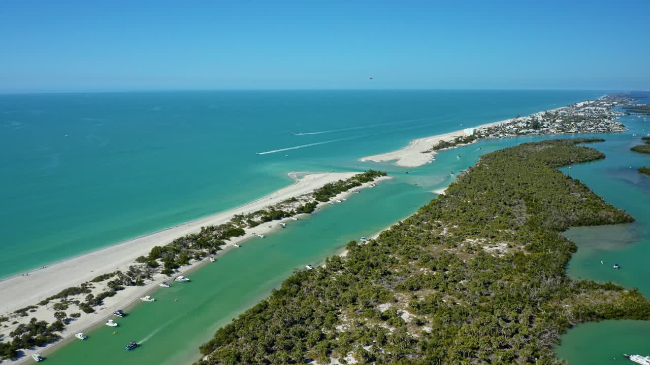 Une crique côtière avec des bateaux ancrés s'étend le long d'une plage de sable et d'un quartier résidentiel, bordée d'une végétation luxuriante sous un ciel bleu clair.