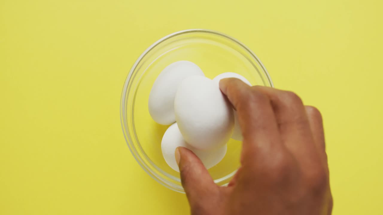 Video of overhead view of glass bowl with eggs on yellow background