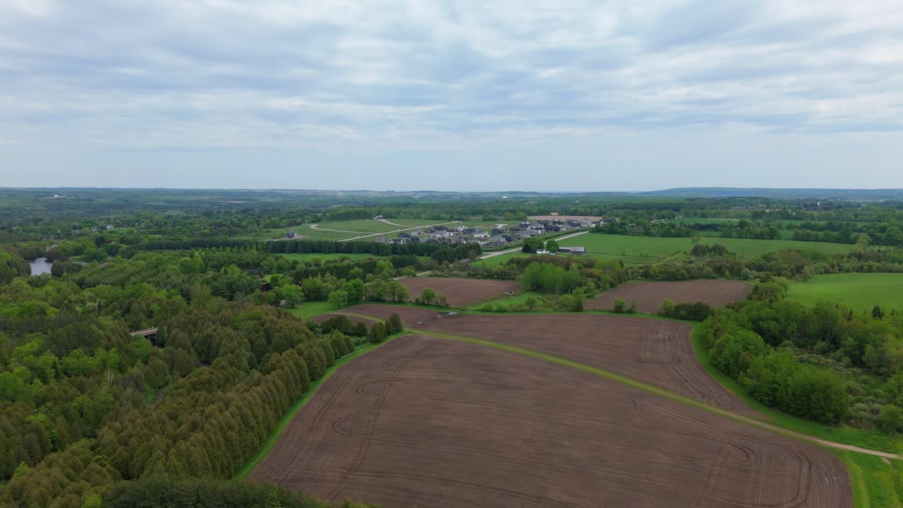 Et droneperspektiv fanger krydsfeltet mellem land- og forstadsliv og viser pløjede, brune marker og frodige, grønne skove i forgrunden med et nyt boligbyggeri i horisonten.