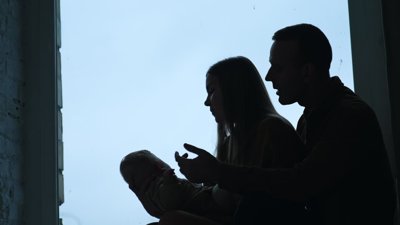 Silhouettes of parents with little baby in hands. Mom and dad sit at window-sill talking to their child.