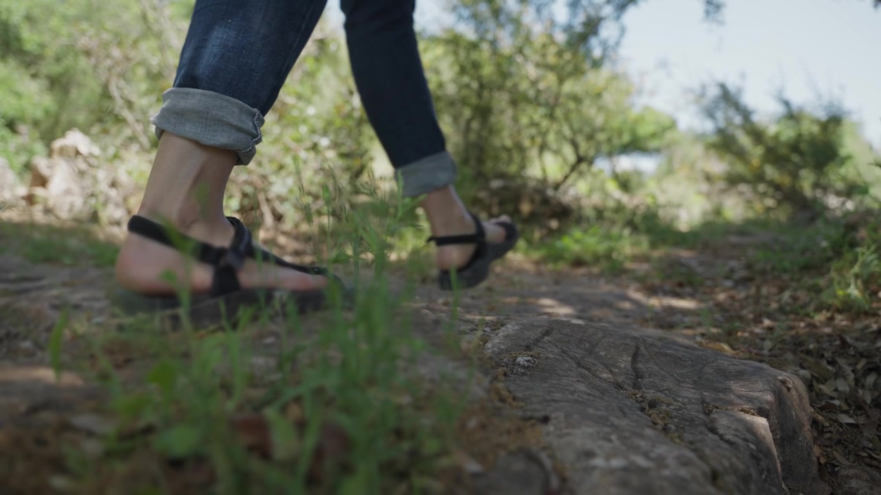 Escursionista donna che cammina con un piccolo zaino e sandali all'aperto su un sentiero naturale. Viaggia in una giornata di sole su un sentiero sterrato. Si gode uno stile di vita sano. Vista dal basso di calzature comode. Erba e legno.