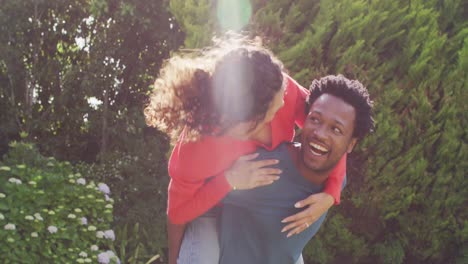 Happy-biracial-man-holding-piggyback-his-fiance-with-engagement-ring-on-hand-in-garden-in-sun