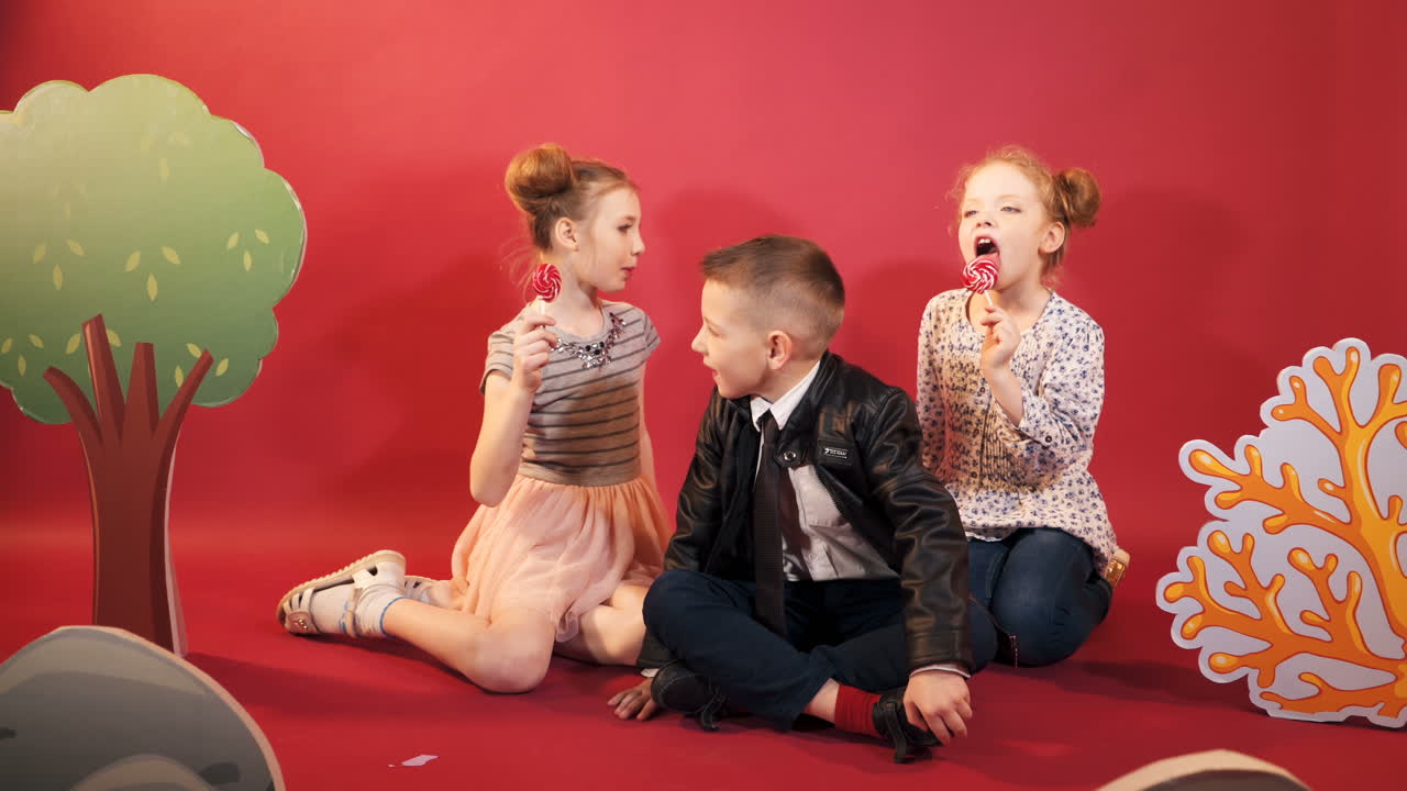 Little children are sitting on the floor on the red background. Two girls are eating lollipops and a cute boy in black suit and a tie sitting in the middle of the girls.