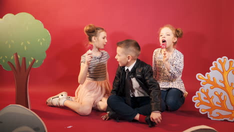 little children are sitting on the floor on the red background. two girls are eating lollipops and a cute boy in black suit and a tie sitting in the middle of the girls.