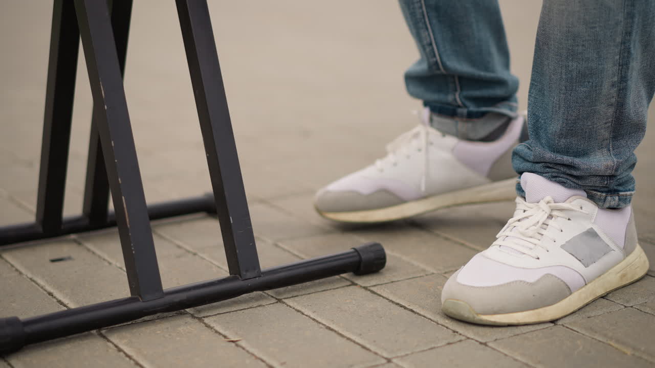 Hvide sneakers på fortov ved side af stander, afslappet DJ-fodarbejde skaber jævn rytme under udendørs gadeoptræden, denimjeans med opsmøg, nærbillede af snørebånd og sål, teksturerede betonfliser, dagtimerne i byen