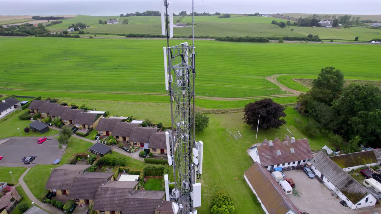 A drone captures a tall communication mast rising above rural Danish houses and green fields on a cloudy day