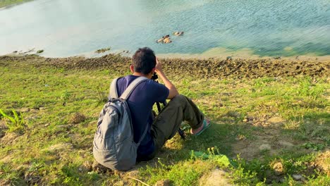 backpack wearing wildlife photographer sitting edge of a lake or river and taking pictures with the help of a tripod