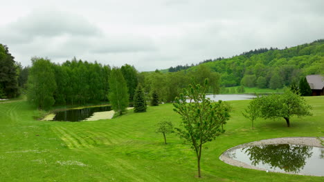 Una-Serena-Vista-Aérea-De-Un-Cuidado-Parque-Con-Pequeños-Estanques,-Exuberante-Vegetación-Y-Colinas-Boscosas-Circundantes