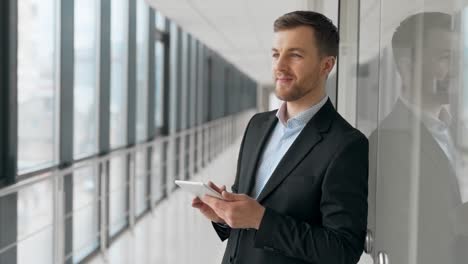 Business-man-with-tablet-smiling-standing-in-a-glass-corridor