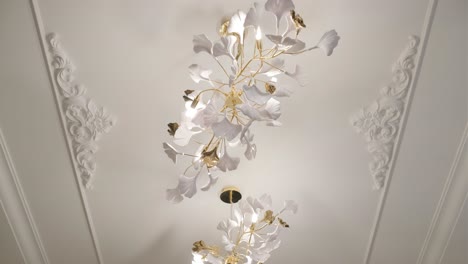 an intricate white and gold ceiling light with floral elements, set against an ornately decorated ceiling with classic moldings