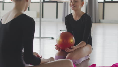 two gymnastic blonde girls talking and laughing while playing with a ball sitting on the floor before starting ballet class