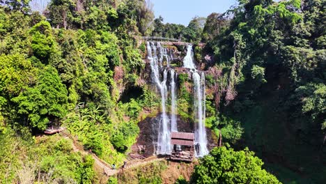 aerial drone orbit reveals tiered tad gneuang waterfall surrounded by lush jungle in champasak province, laos, with misty cascades flowing beside a traditional wooden pavilion built on the cliffside