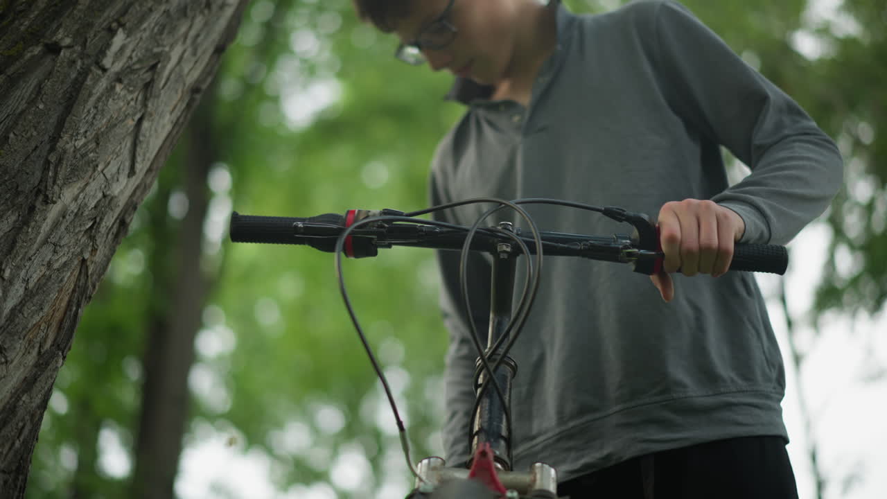 niño con gafas agarra firmemente el manillar de una bicicleta estacionada cerca de un árbol en un campo cubierto de hierba, está comprobando la funcionalidad de los frenos mientras está de pie, su cara está borrosa en el fondo