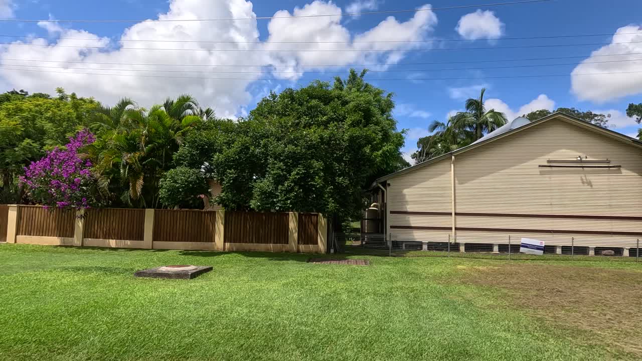 A vehicle moves past suburban houses and greenery under a bright sky in Mullumbimby, Australia. The scene captures a peaceful neighborhood
