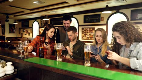 young people are checking smartphones while sitting at bar counter together. they are showing each other screens, laughing and talking. modern technologies communication concept.