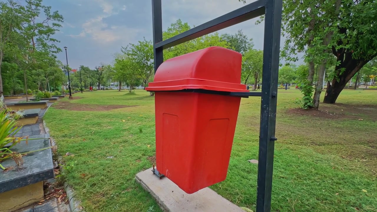 Tomada de câmera se aproximando de uma lixeira vermelha instalada em um parque público, cercada por gramados verdes e árvores, enfatizando a limpeza e a manutenção urbana.