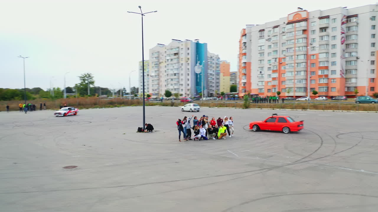 Teenage group of dancers moving in the middle of the square. Race car drifting around the dancers. Blocks of flats at backdrop.