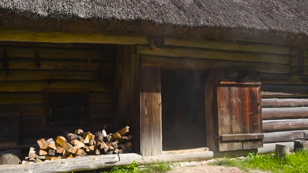 Low angle slow pan of traditional smoke sauna, ancient log house. Barn dwelling facade with thatched roof and stacked firewood. Open chamber wooden door releasing stream of smoke into the summer air