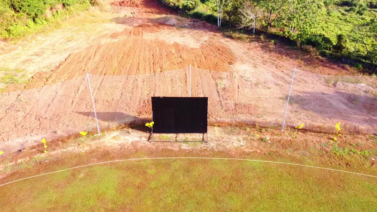 Aerial drone shot zooming into a large black sightscreen on a cricket field, featuring a lush green outfield bordering a red clay construction zone surrounded by tropical forest
