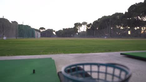 área-De-Práctica-De-Un-Campo-De-Golf,-La-Cámara-Baja-Lentamente-Y-Cambia-El-Enfoque-A-Un-Cubo-De-Pelota-Vacío-Que-Se-Encuentra-Junto-A-Una-Alfombra