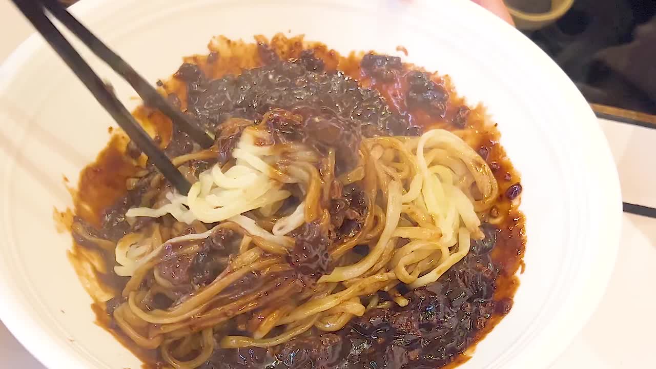 A detailed view of noodles being mixed with a rich, dark sauce using chopsticks.