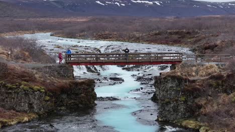 A man stands on the precipice of a bridge, holding a drone as he explores the hidden treasures of the land and unveils a mesmerizing display of natural marvels - the majestic Bruararfoss waterfall.