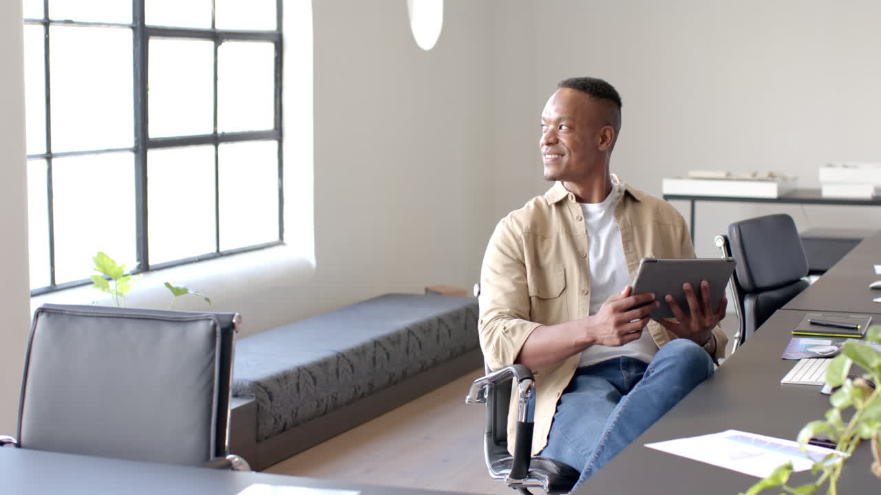 Smiling businessman holding tablet and sitting in modern office workspace