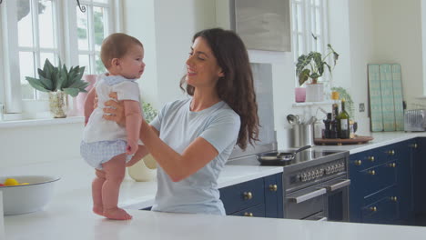 Loving-Mother-Playing-With-Baby-Son-At-Home-Standing-On-Kitchen-Counter