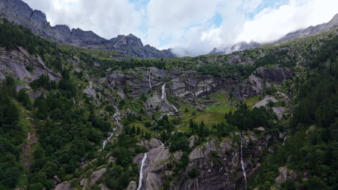 Aerial drone shot pulling back and tilting upward from a long waterfall on rocky cliffs, revealing mountains and cloudy sky in the Swiss Alps