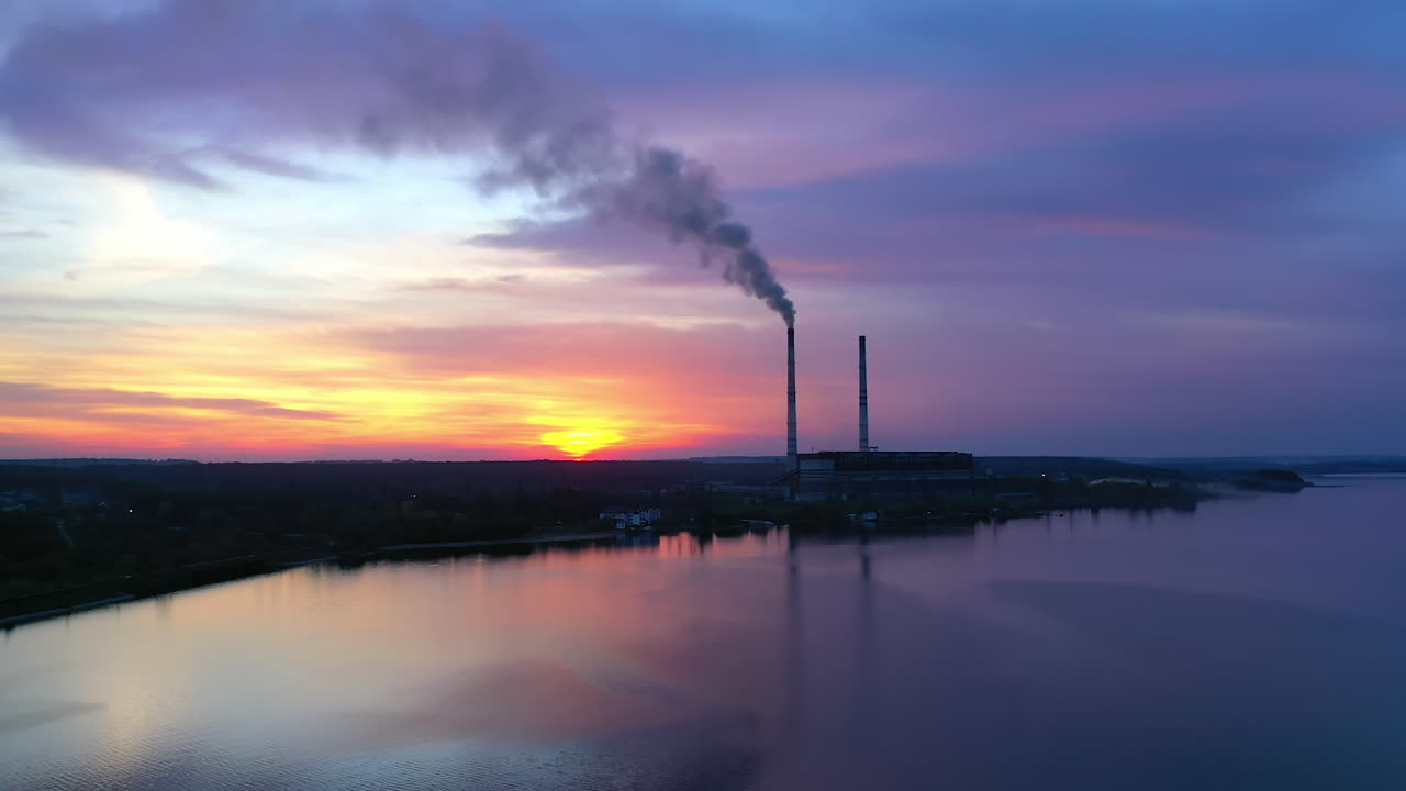 Factory near the river in the evening. Dirty smoke goes from industrial plant at sunset. Calm river on chemical manufacturing background. Air pollution. Aerial view.
