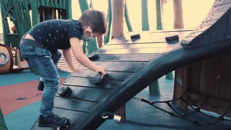 cute preschooler with face mask climbing on the wooden wall in the park