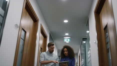 Multiracial-business-team-talking-finance-plan-using-tablet-in-office-hallway.