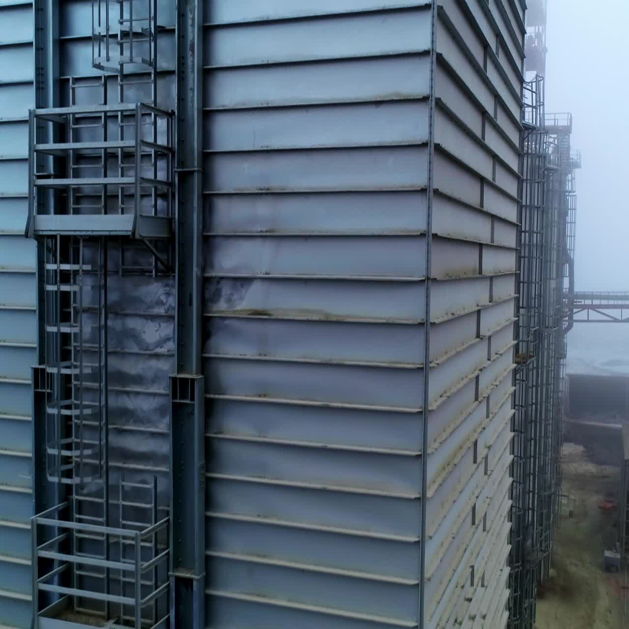 High metal rectangular premise emitting thick grey smoke. Going up along the one of the constructions at modern silo complex