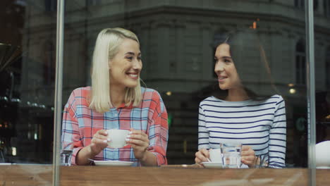 Retrato-De-Dos-Amigas-Tomando-Café-Y-Sonriendo-A-La-Cámara-Mientras-Están-Sentadas-En-Un-Café