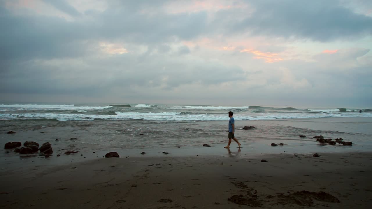 A young man walks along Echo Beach in Bali at sunset, showcasing a serene coastal scene with soft evening light, gentle waves, and a peaceful, reflective atmosphere.