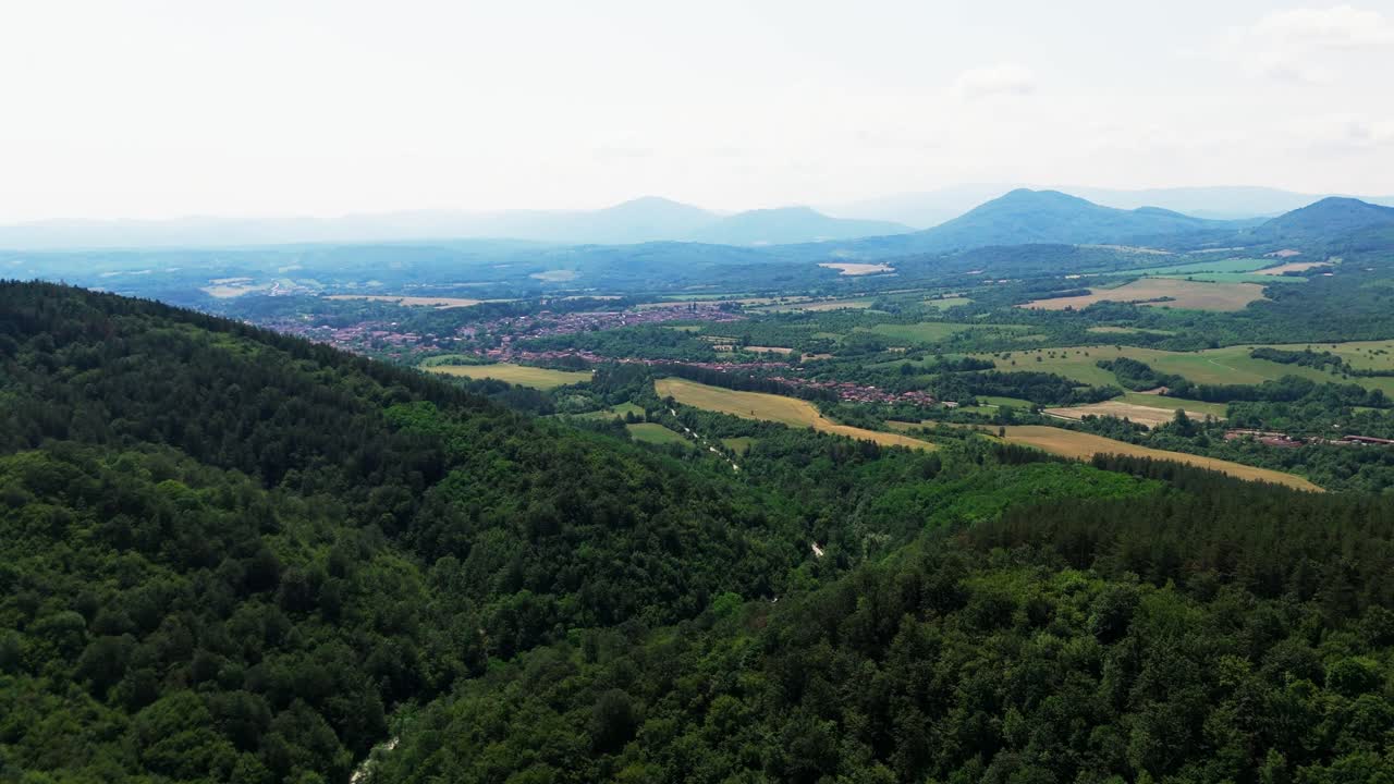 Ripresa con drone che rivela un vasto paesaggio balcanico con valli, colline, villaggi, campi e montagne velate all'orizzonte.