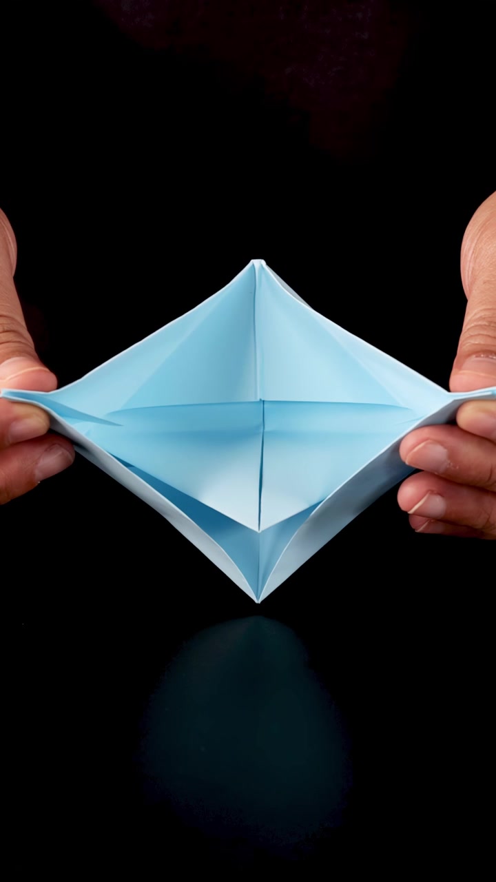 Manos de una persona doblan cuidadosamente papel azul para formar un barco de origami sobre un fondo oscuro.