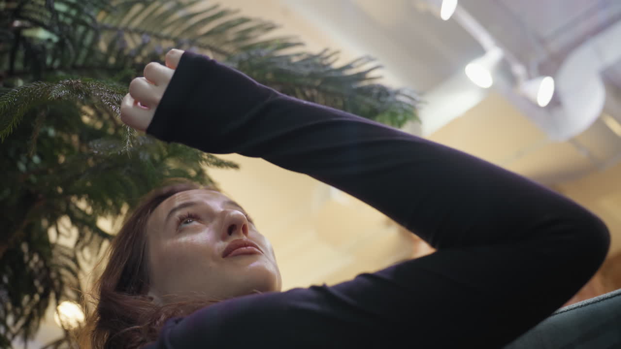 Closeup of woman in black long-sleeve top looking upward while gently reaching to touch pine leaf in brightly lit indoor setting with soft light reflections and exposed brick wall in background