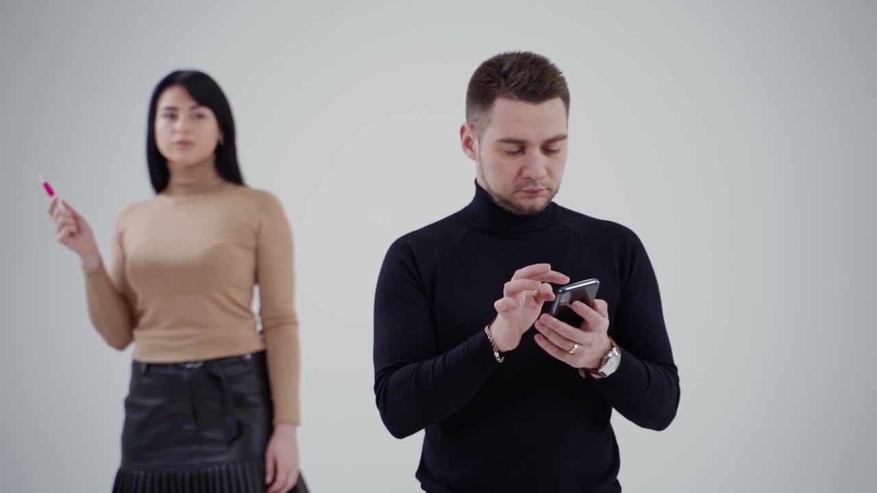 Young man in black sweater clicking the mobile phone. Attractive sad woman standing behind the man and smoking on the studio background.